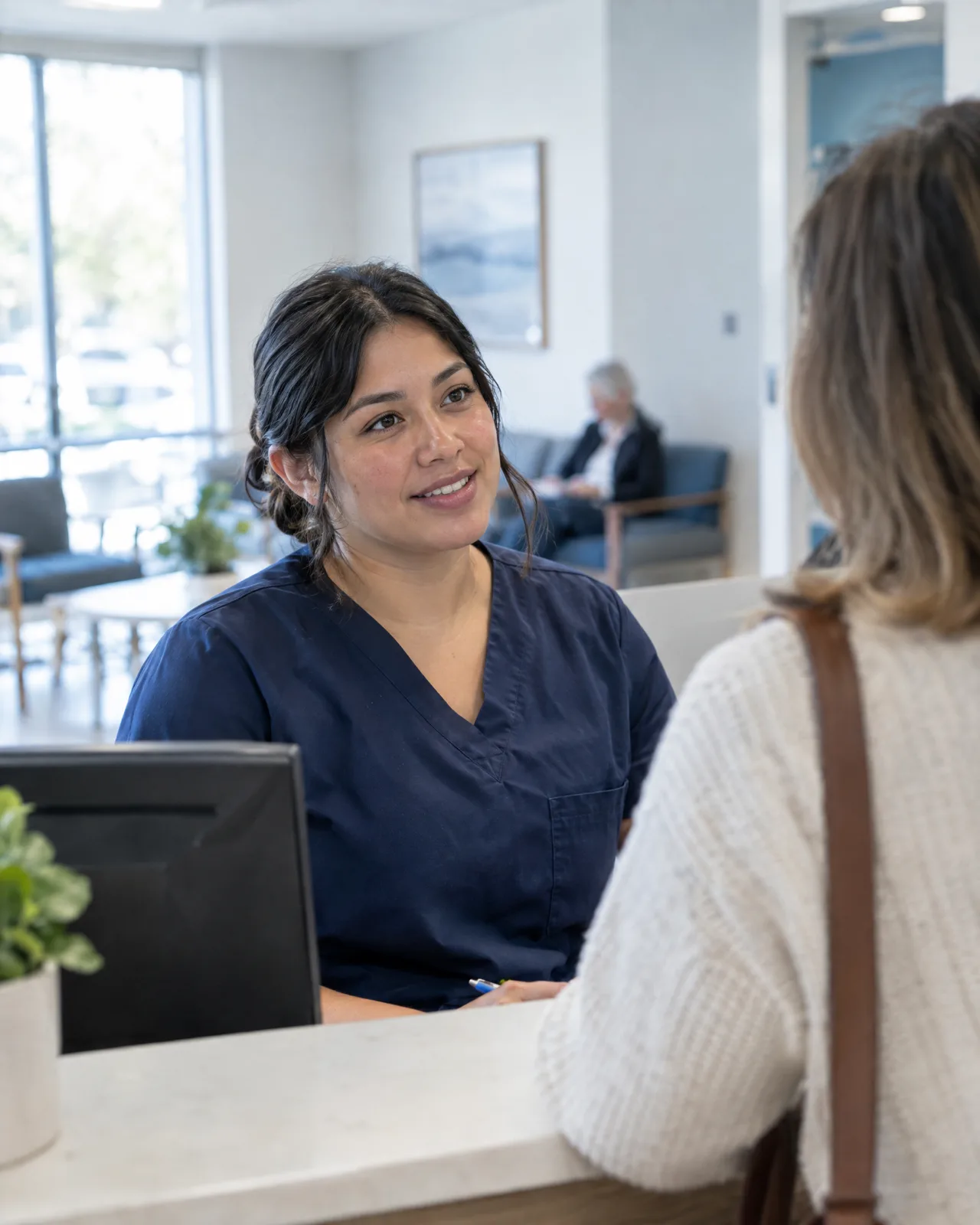 A medical assistant in scrubs talking with a patient at a clinic front desk.